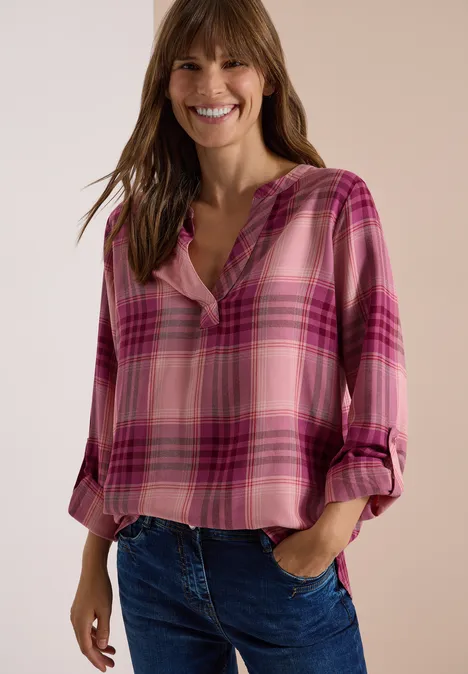 Eine lächelnde Frau trägt eine pink karierte Bluse und Jeans vor hellem Hintergrund.