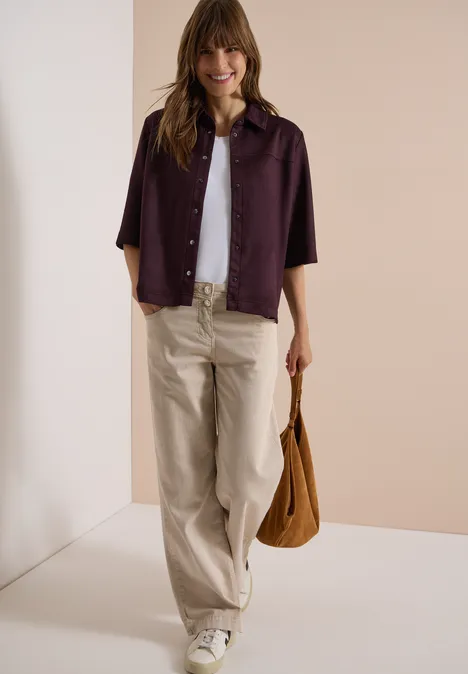 Frau trägt weiße Bluse, lila Hemdjacke, beige Hose und braune Tasche vor neutralem Hintergrund.