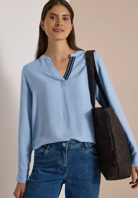 Langarm Tunika mit Split Neck und Tape blouse blue melange Eine lächelnde Frau trägt eine hellblaue Bluse, Jeans und eine schwarze Tasche.