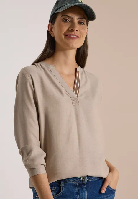 Langarm Tunika mit Split Neck und Tape creamy mocha melange Frau in beiger Bluse, Jeans und grüner Kappe, lächelnd, mit Händen in den Hosentaschen.