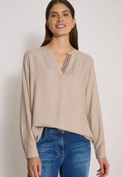 Langarm Tunika mit Split Neck und Tape creamy mocha melange Frau in beige Bluse mit V-Ausschnitt und blauen Jeans vor hellem Hintergrund lächelnd.