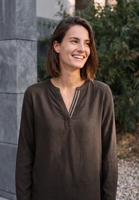 Une femme souriante en chemise marron se tient devant un mur de pierre et des plantes.