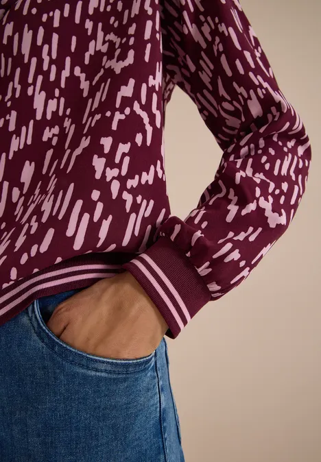 Nahaufnahme: Person in blauen Jeans, Hand in Tasche, burgunderfarbener Pullover mit weißem Muster.