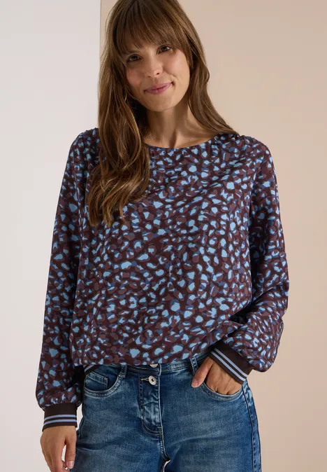 Vrouw in bruine blouse met blauw patroon en jeans tegen een neutrale achtergrond.