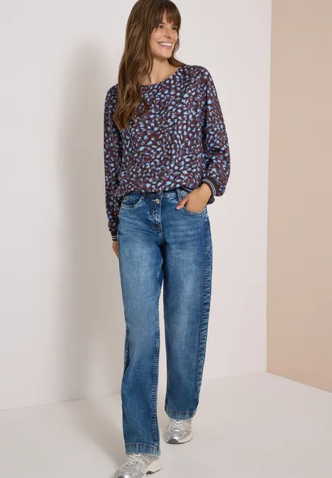 Vrouw in een blouse met print, rechte jeans en sneakers, staat ontspannen poseert en lacht.