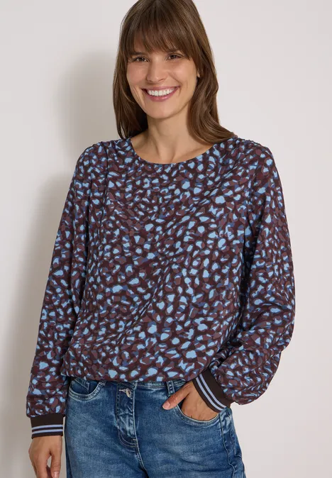 Vrouw lacht in bruin-blauw dessin top en blauwe jeans tegen een lichte achtergrond