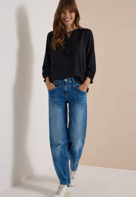 Frau lehnt an Wand, trägt schwarze Bluse, lockere blaue Jeans und weiße Sneaker.