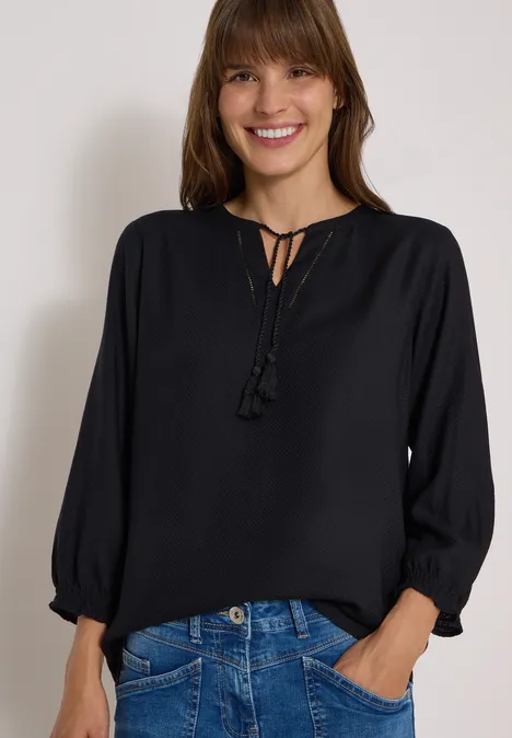 Junge Frau in schwarzer Bluse mit Quasten und blauen Jeans, lächelnd mit Händen in den Taschen.