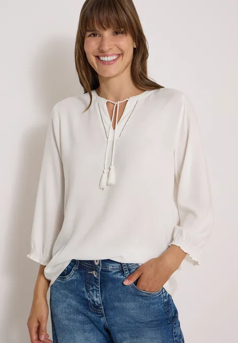 Eine lächelnde Frau trägt eine weiße Bluse mit Quasten und blaue Jeans.