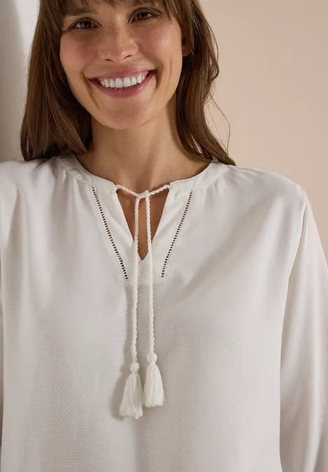 Eine lächelnde Frau trägt eine weiße Bluse mit Quasten und Bindebändern.
