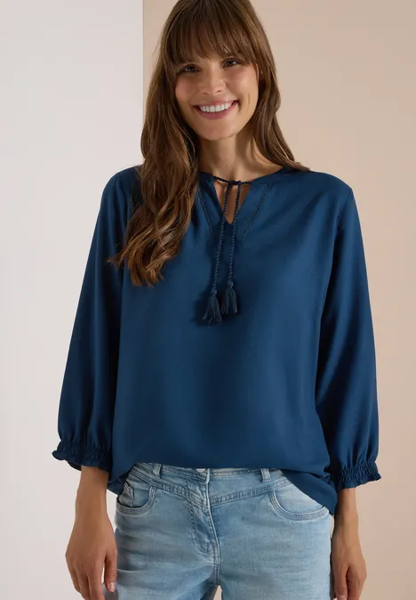 Frau lächelt in dunkelblauer Bluse mit Quasten und hellen Jeans.