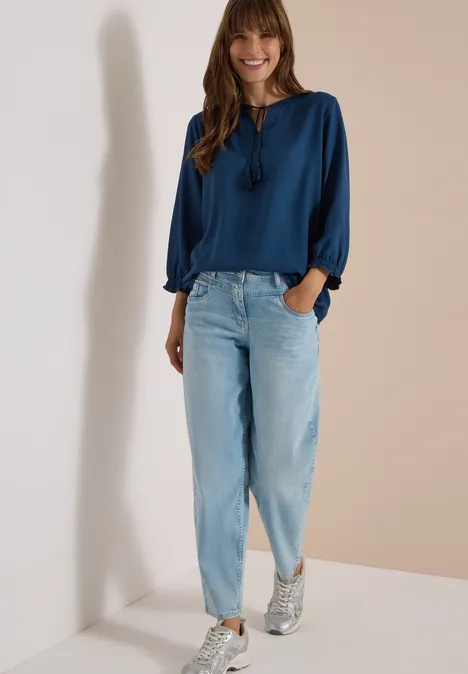 Eine lächelnde Frau lehnt an einer weißen Wand, trägt eine blaue Bluse, helle Jeans und Sneaker.