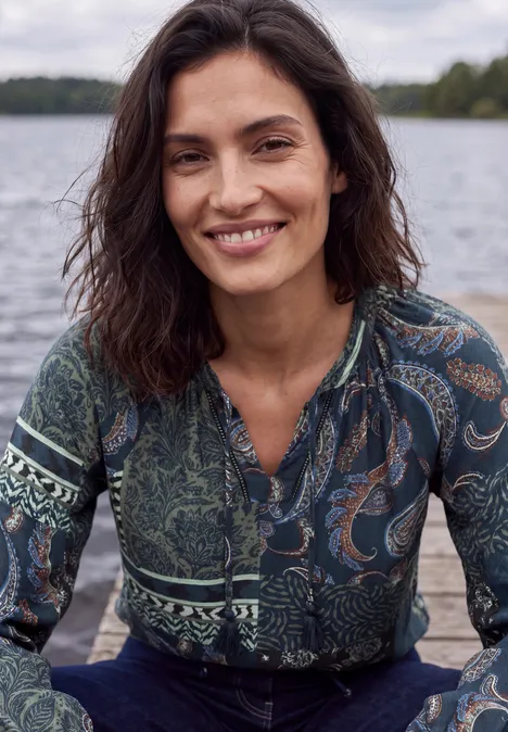 Glimlachende vrouw met donker haar en een patroonblouse, zittend aan de oever van een meer.