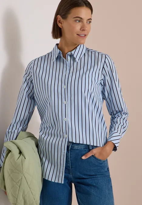 Chemisier long à rayures blouse blue Femme en chemise rayée bleue, jean et une veste matelassée verte claire à la main.