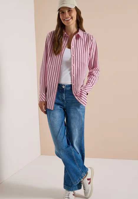 Chemisier long à rayures mellow rose Femme porte une chemise rose et blanche à rayures, t-shirt blanc, jean bleu et baskets blanches sur fond neutre.