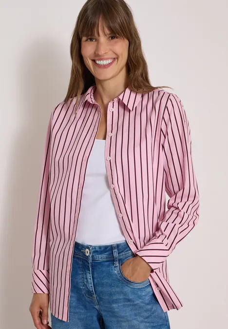 Chemisier long à rayures mellow rose Une femme sourit, portant une chemise rose à rayures blanches sur un débardeur blanc et un jean.