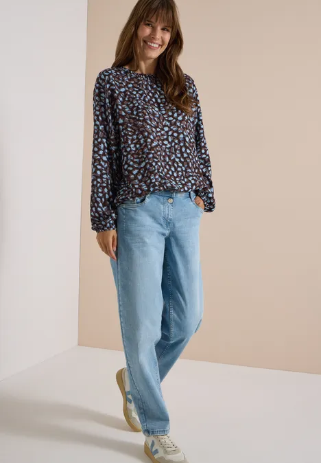 Een lachende vrouw in een kleurrijk blouse en lichte spijkerbroek staat nonchalant voor een neutrale achtergrond.