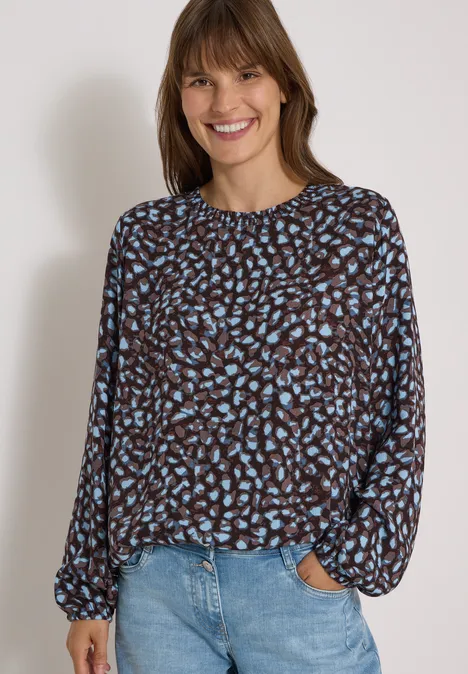 Jonge vrouw met een gebloemde blouse en jeans, glimlachend voor een lichte achtergrond.