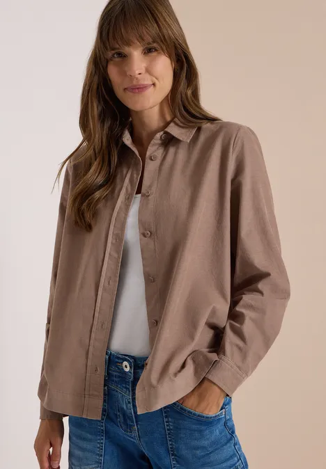 Chemisier à manches longues en velours côtelé earthy taupe Femme portant une chemise marron, un débardeur blanc et un jean, les mains dans les poches, souriante.