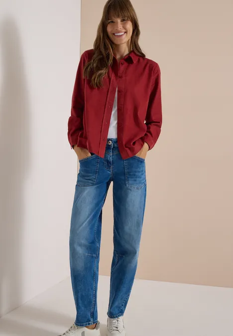 Chemisier à manches longues en velours côtelé russet red Femme souriante en veste rouge, t-shirt blanc et jean bleu, appuyée contre un mur, mains dans les poches.