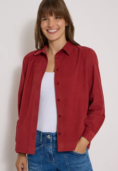 Chemisier à manches longues en velours côtelé russet red Une femme souriante porte une chemise rouge sur un débardeur blanc et un jean bleu.