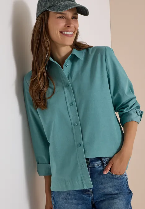 Chemisier à manches longues en velours côtelé mineral blue Une femme souriante appuyée contre un mur, portant une chemise verte, un jean et une casquette.