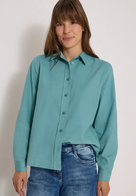 Chemisier à manches longues en velours côtelé mineral blue Une femme souriante porte une chemise turquoise à manches longues et un jean devant un mur clair.