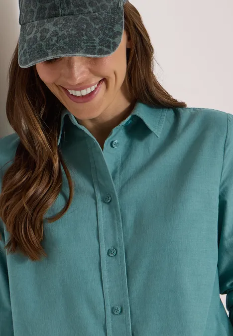 Chemisier à manches longues en velours côtelé mineral blue Femme en chemise verte et casquette motif sourit vers le bas.