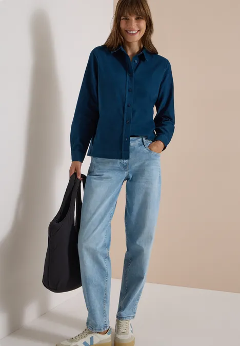 Chemisier à manches longues en velours côtelé moonlight blue Femme en chemise bleu foncé, jean clair et baskets claires avec un sac noir, posant devant un mur neutre.