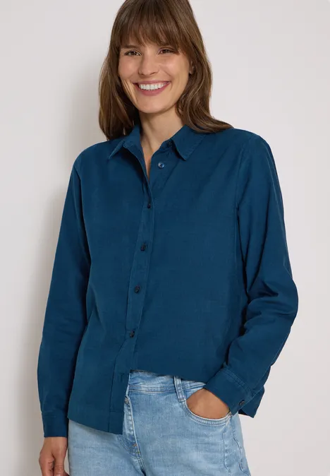 Chemisier à manches longues en velours côtelé moonlight blue Femme souriante portant une chemise bleue et un jean, main dans la poche, fond clair.