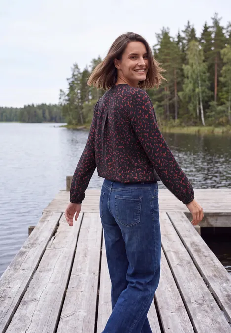 Tunique à manches longues, à col fendu et imprimé urban dark blue Une femme en chemisier fleuri et jean sourit sur un ponton au bord d'un lac, forêt en arrière-plan.