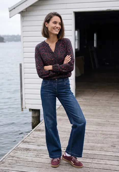 Tunique à manches longues, à col fendu et imprimé urban dark blue Une femme souriante en jean et chemise, bras croisés, se tient sur un ponton devant un abri à bateaux blanc.
