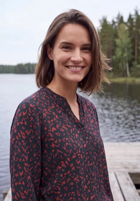 Tunique à manches longues, à col fendu et imprimé urban dark blue Une femme souriante se tient sur un quai en bois au bord d’un lac, avec une forêt en arrière-plan.