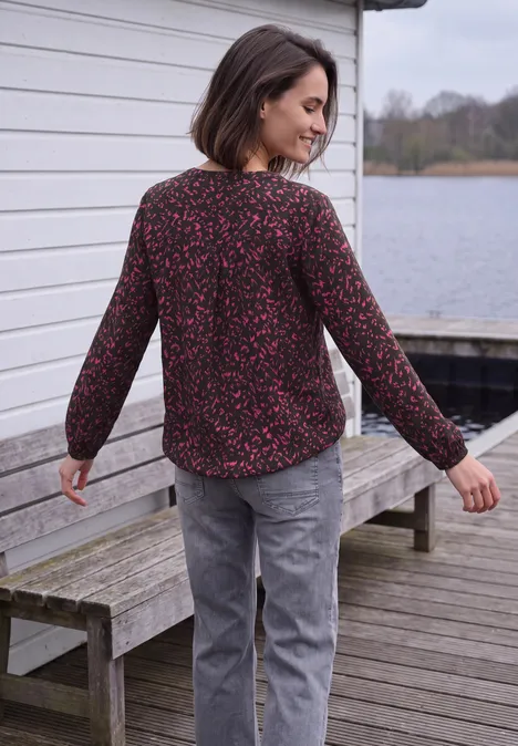Frau steht auf einem Holzsteg am See und schaut über die Schulter, sie trägt geblümte Bluse und graue Hose.