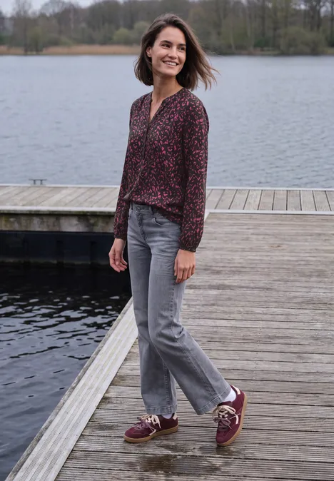 Eine Frau in gemustertem Oberteil, grauen Jeans und roten Sneakern steht auf einem Steg am See.