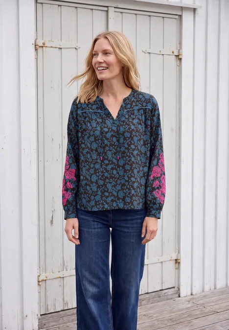 Eine lächelnde Frau in blau gemusterter Bluse mit pinken Ärmeln und Jeans vor einer weißen Holzwand.