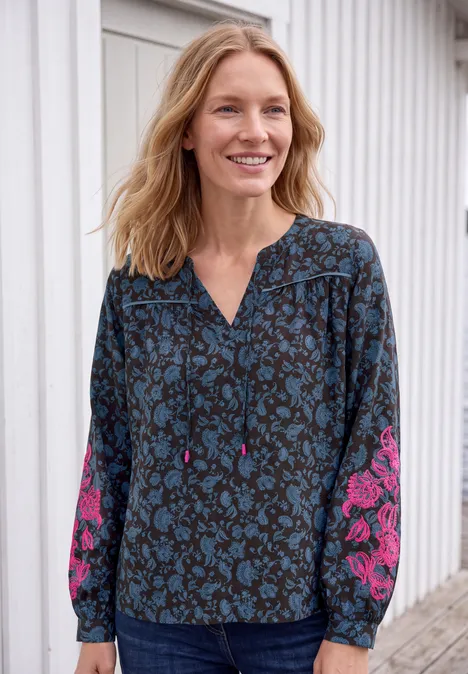Frau lächelt vor weißer Wand, trägt Bluse mit blauem und pinkem Blumenmuster.