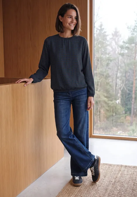 Langarm Bluse mit Rundhals und Print urban dark blue Eine lächelnde Frau in Jeans und dunklem Pullover lehnt an einer Holztheke und schaut aus dem Fenster.