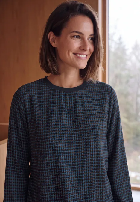 Langarm Bluse mit Rundhals und Print urban dark blue Eine lächelnde Frau in kariertem, dunklem Oberteil vor einem Fenster mit unscharfem Hintergrund.
