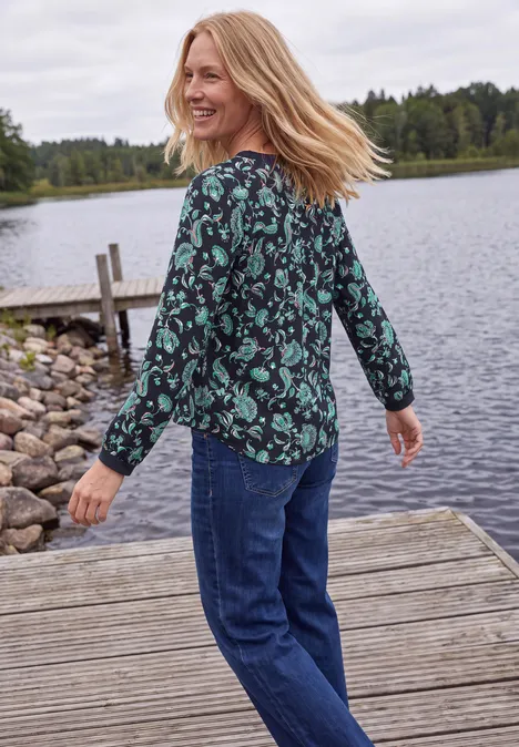 Chemisier à manches longues, à col fendu et imprimé urban dark blue Femme souriante regarde par-dessus l'épaule sur un ponton en bois au bord d'un lac, blouse verte à fleurs et jean.