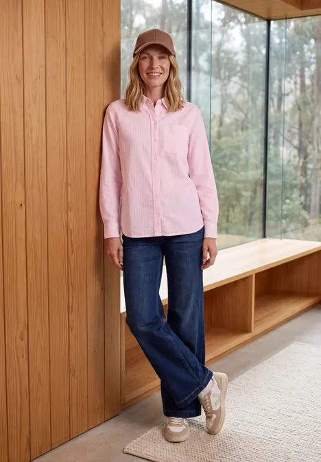 Langarm Hemdbluse mit Streifen mellow rose Frau in rosa Hemd, blauen Jeans und Mütze lehnt lächelnd an einer Holzverkleidung neben großen Fenstern.