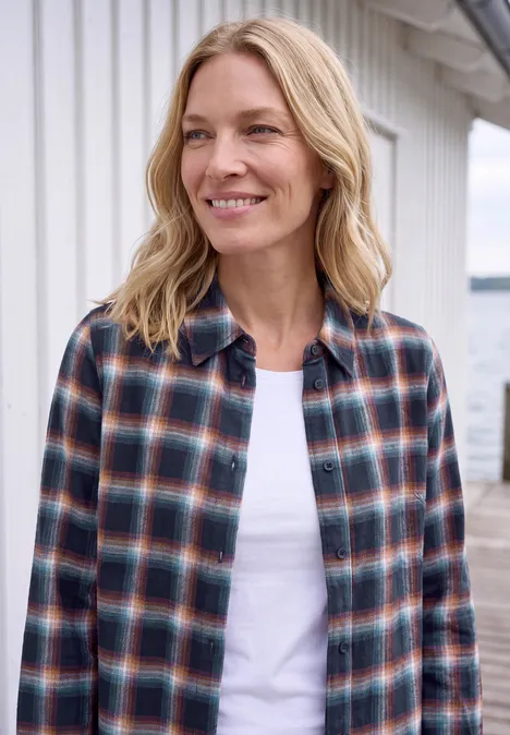 Chemisier en flanelle à manches longues et à carreaux urban dark blue Une femme blonde souriante porte une chemise à carreaux sur un t-shirt blanc devant une maison en bois claire.