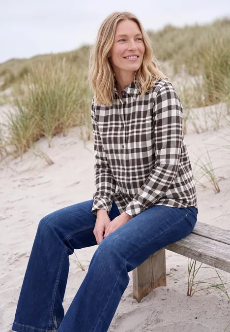 Eine lächelnde Frau sitzt auf einer Holzbank am Strand mit kariertem Hemd und Jeans.