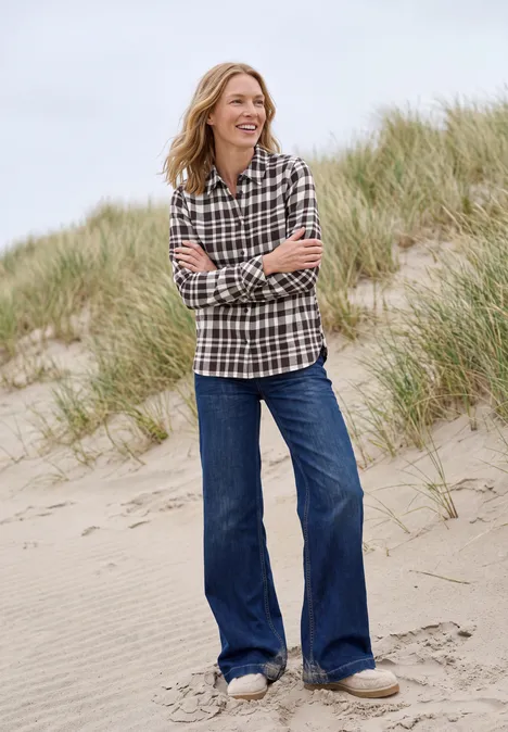 Eine lächelnde Frau in kariertem Hemd und weitem Jeans steht auf einer Sanddüne mit Strandgras.
