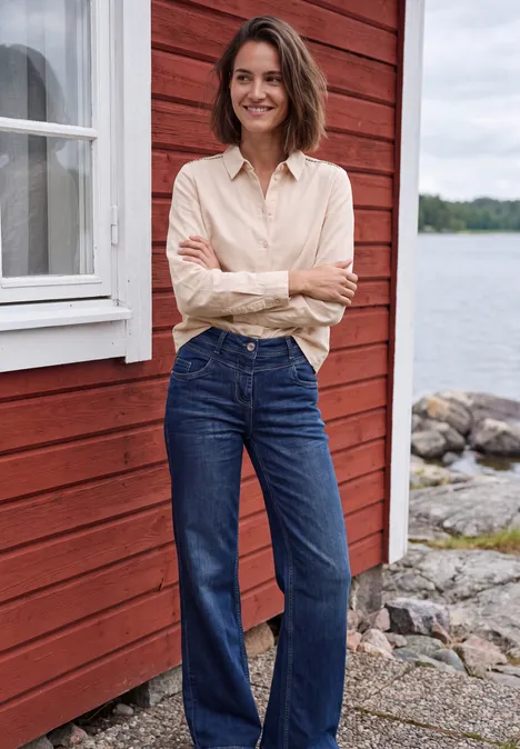 Chemisier à manches longues en velours côtelé uni cashmere beige Femme en jean et chemise claire devant une maison en bois rouge au bord du lac, souriante.