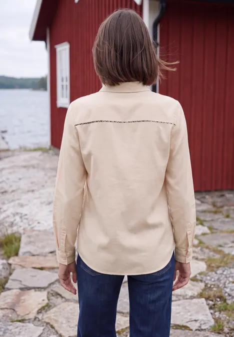 Chemisier à manches longues en velours côtelé uni cashmere beige Une personne en veste beige et jeans vue de dos devant une maison rouge au bord de l'eau.