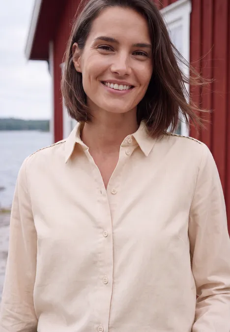 Chemisier à manches longues en velours côtelé uni cashmere beige Une femme souriante en chemise claire devant l'eau et une maison en bois rouge.