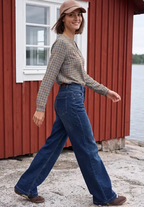 Langarm Tunika mit Split Neck und Print mocha brown Frau in Kappe und kariertem Hemd, weiten Jeans, die vor einem roten Haus am Wasser spaziert.