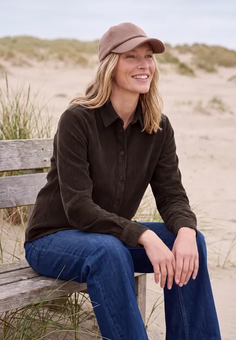 Eine lächelnde Frau in brauner Jacke und Cap sitzt entspannt auf einer Bank am Strand mit Dünen im Hintergrund.