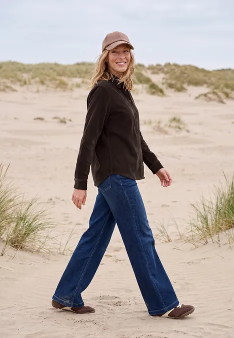 Frau in schwarzer Jacke, Jeans und Mütze spaziert lächelnd über Sanddünen am Strand.